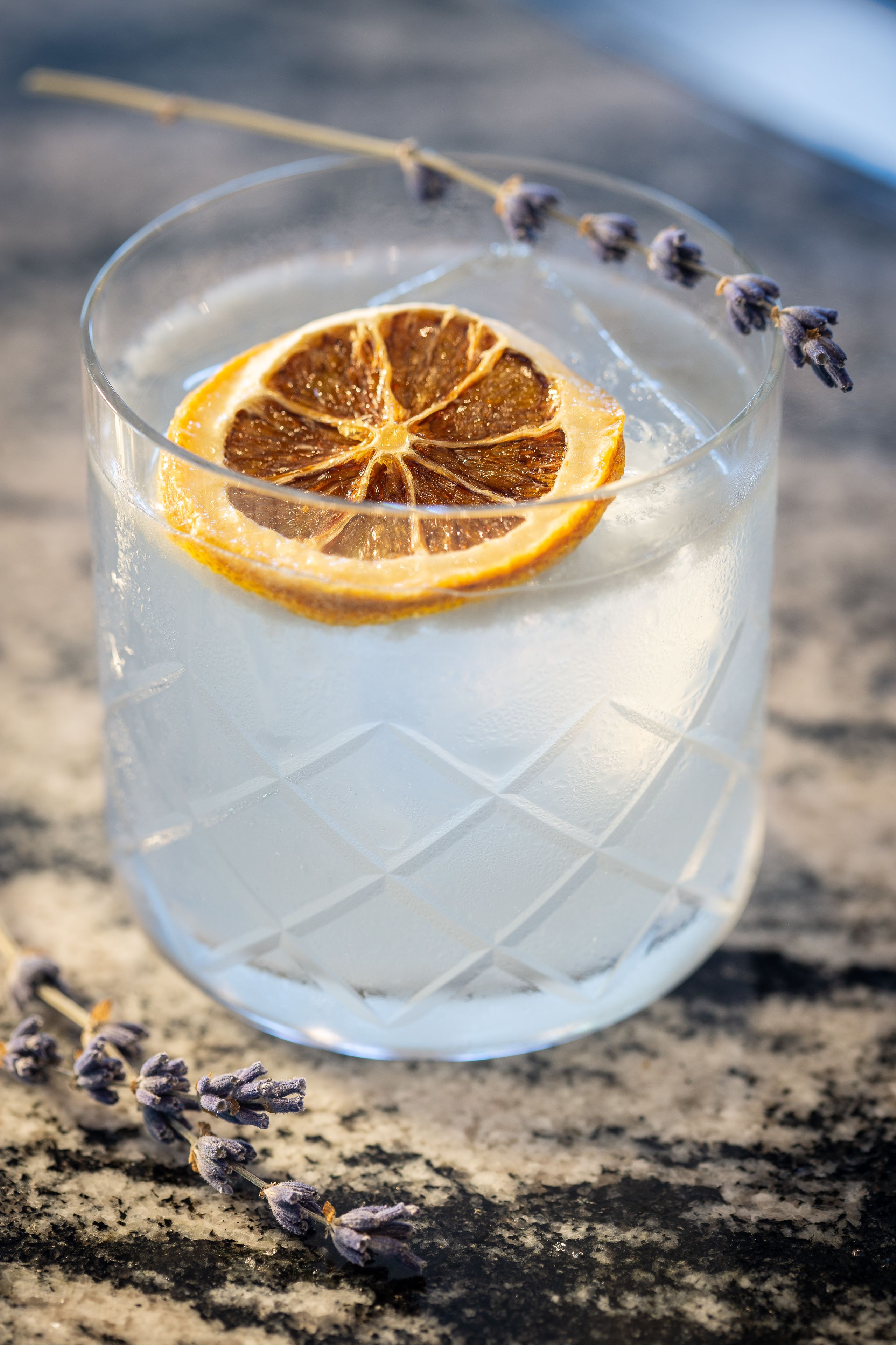 A clear glass with ice and a dried orange slice, placed on a textured surface with lavender sprigs.