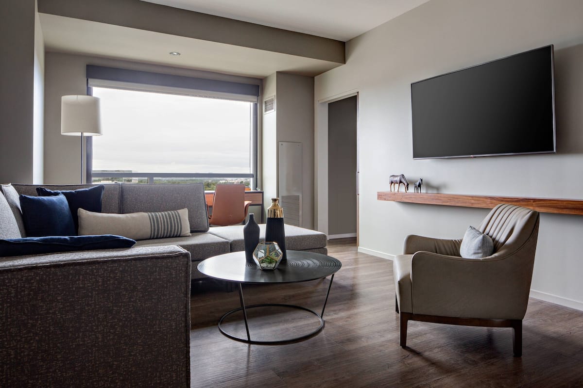 A modern living room with a sofa, armchair, and round coffee table. A large flat-screen TV is mounted on the wall, and a window provides a view of the city.