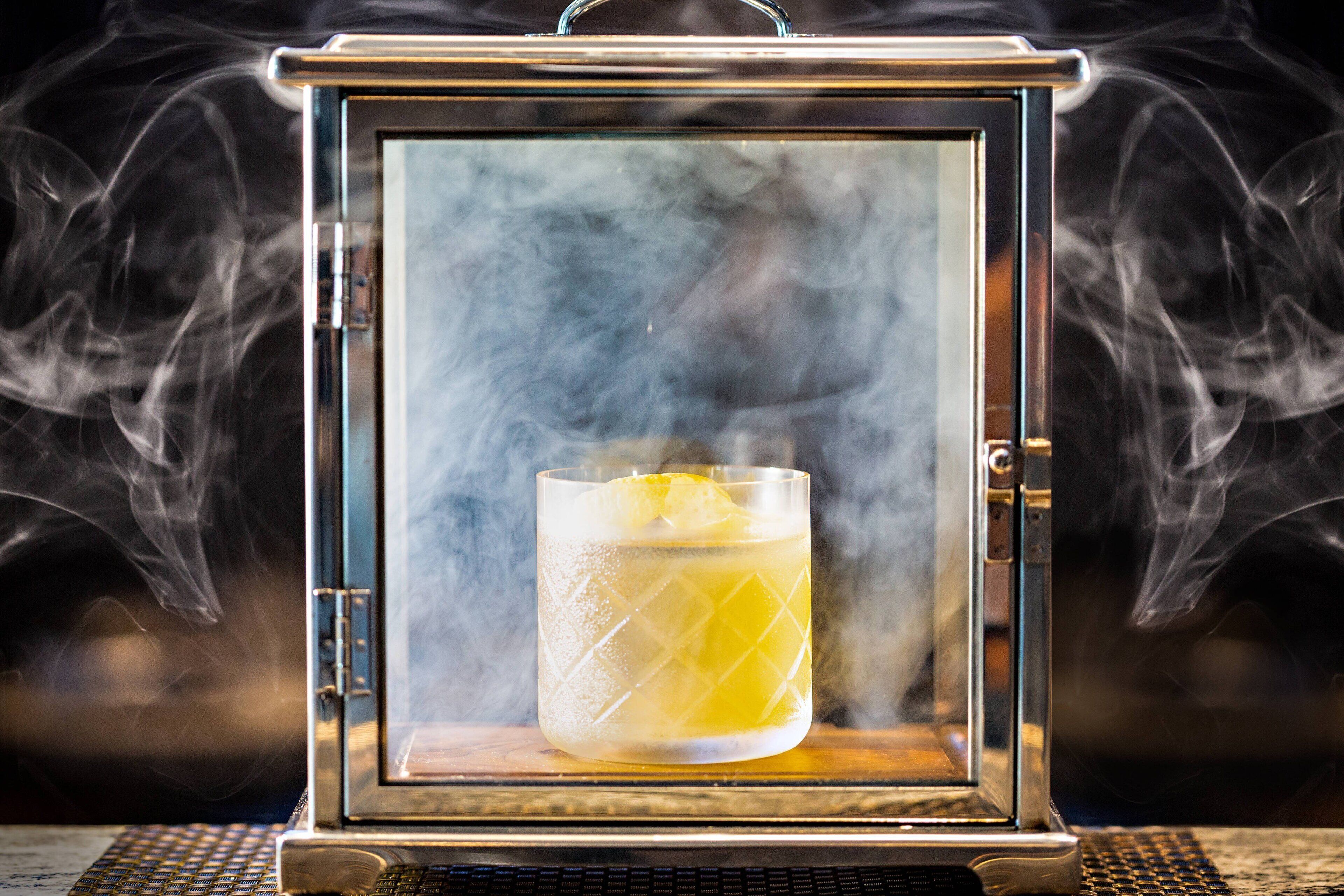 Un vaso de una bebida amarilla con una rodaja de limón dentro de un recipiente antiguo lleno de humo.