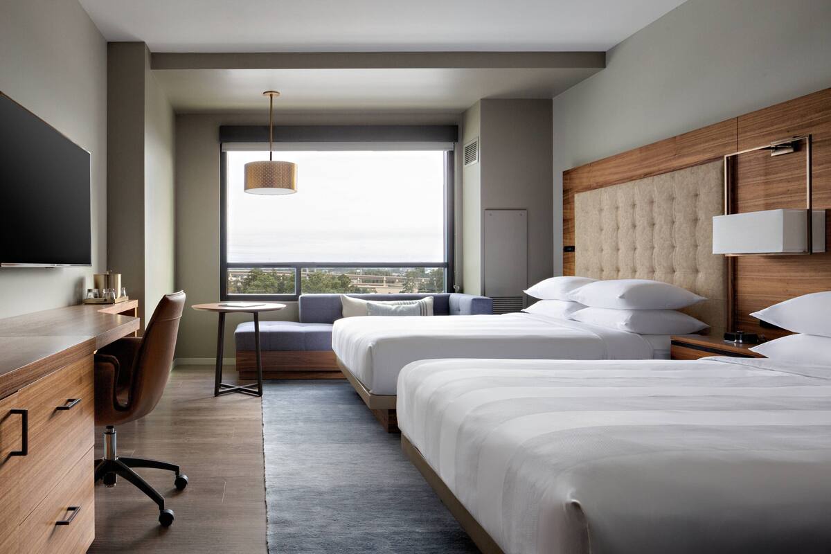 A modern hotel room with two beds, a desk, a chair, and a large window offering a city view.