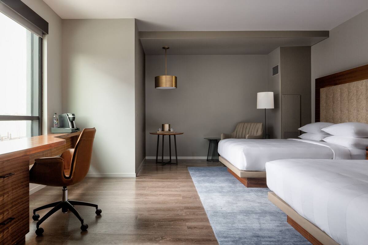 A hotel room with a wooden desk, a chair, a bed, a bedside table, a lamp, and a window.