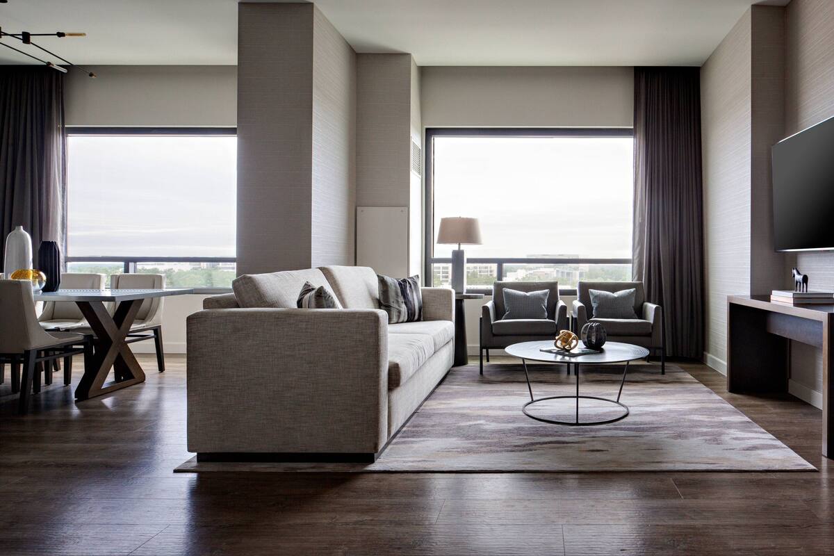 A modern hotel room with a sofa, armchair, and a dining area with a table and chairs.