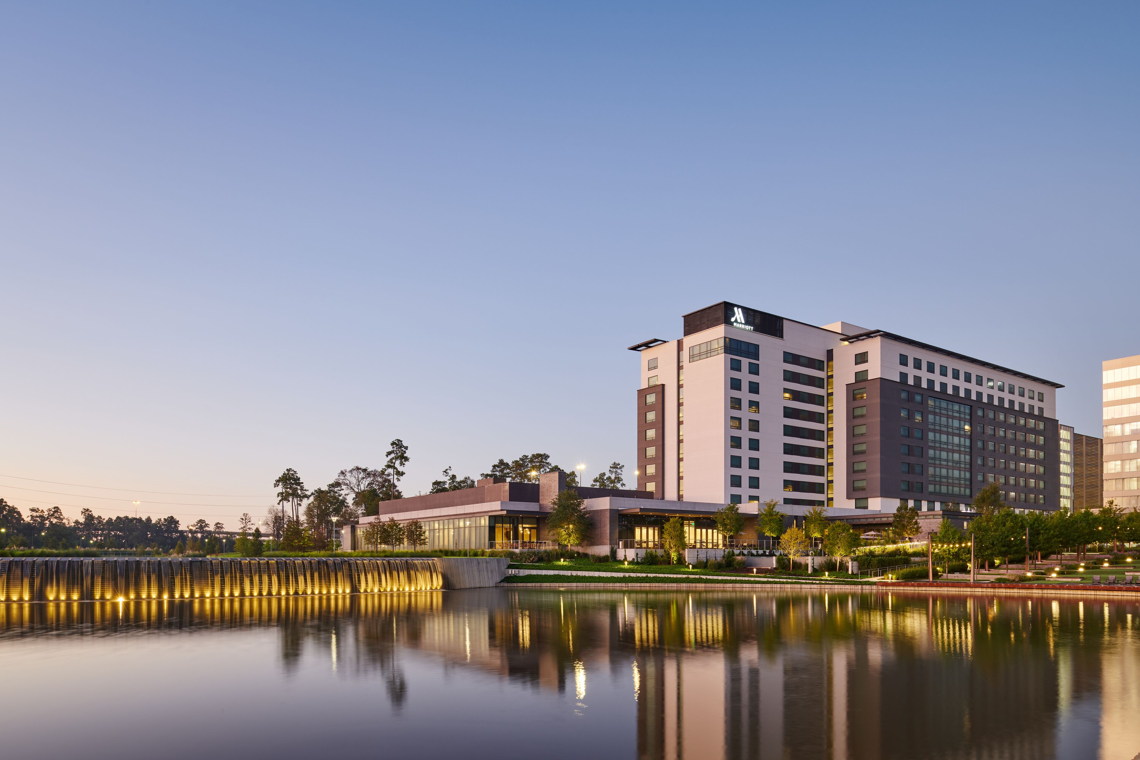 A modern hotel with a reflective water feature in front, surrounded by greenery and trees.