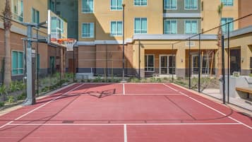 An outdoor basketball court adjacent to a multi-story building with a red surface and a basketball hoop.