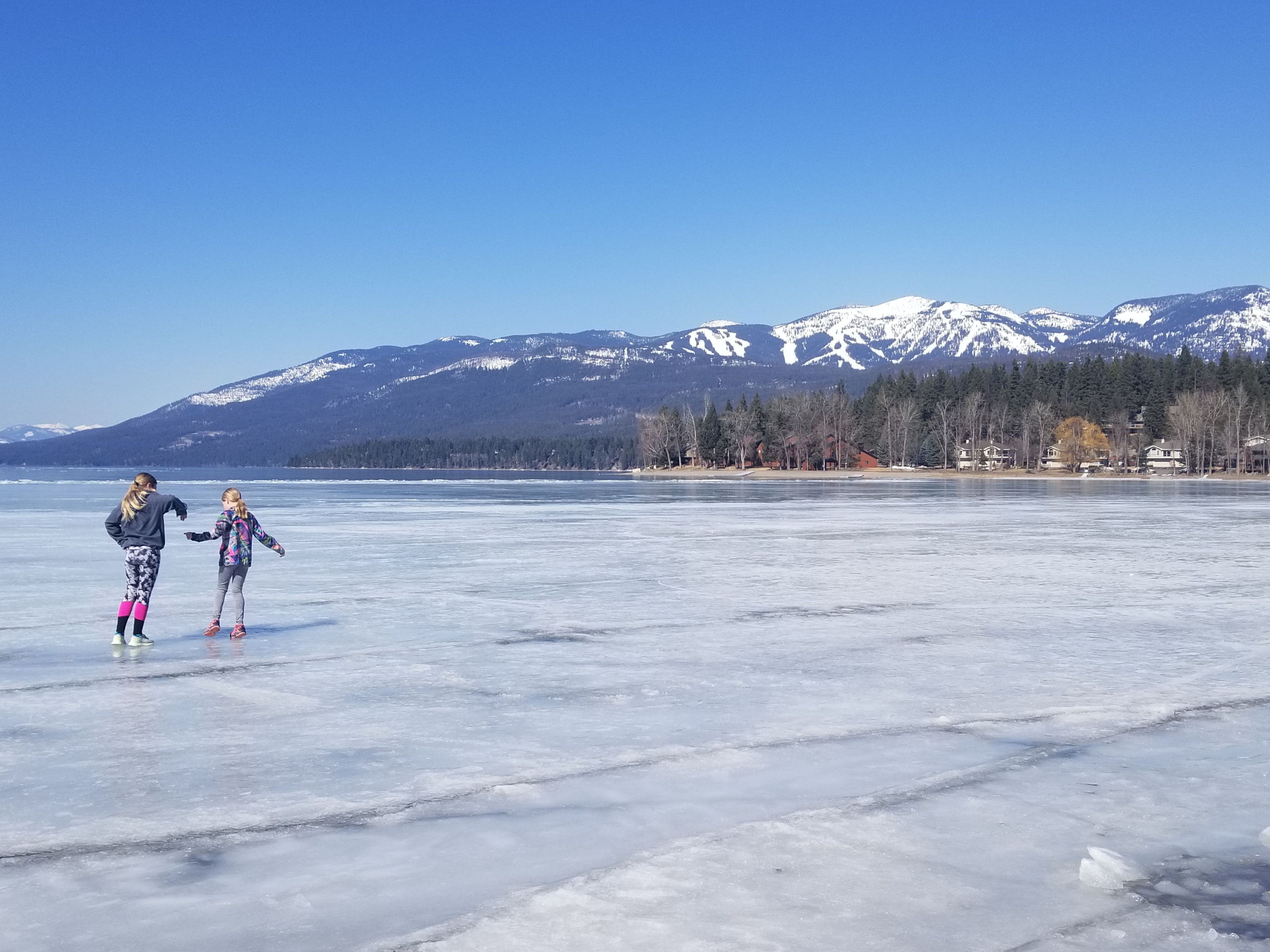 A frozen lake with two people ice skating, surrounded by mountains and houses.