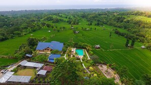 An aerial view of a lush green landscape with terraced fields, a swimming pool, and several buildings with blue roofs. - Ume Cantik Villa (Sukasada)