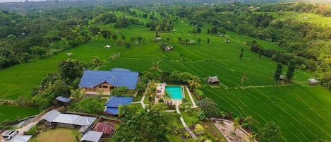 An aerial view of a lush green landscape with terraced fields, a swimming pool, and several buildings with blue roofs.