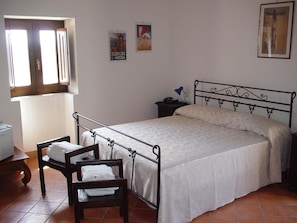 Une chambre avec un lit en métal, une table de chevet avec une lampe, une chaise avec une serviette et une fenêtre avec des rideaux.
