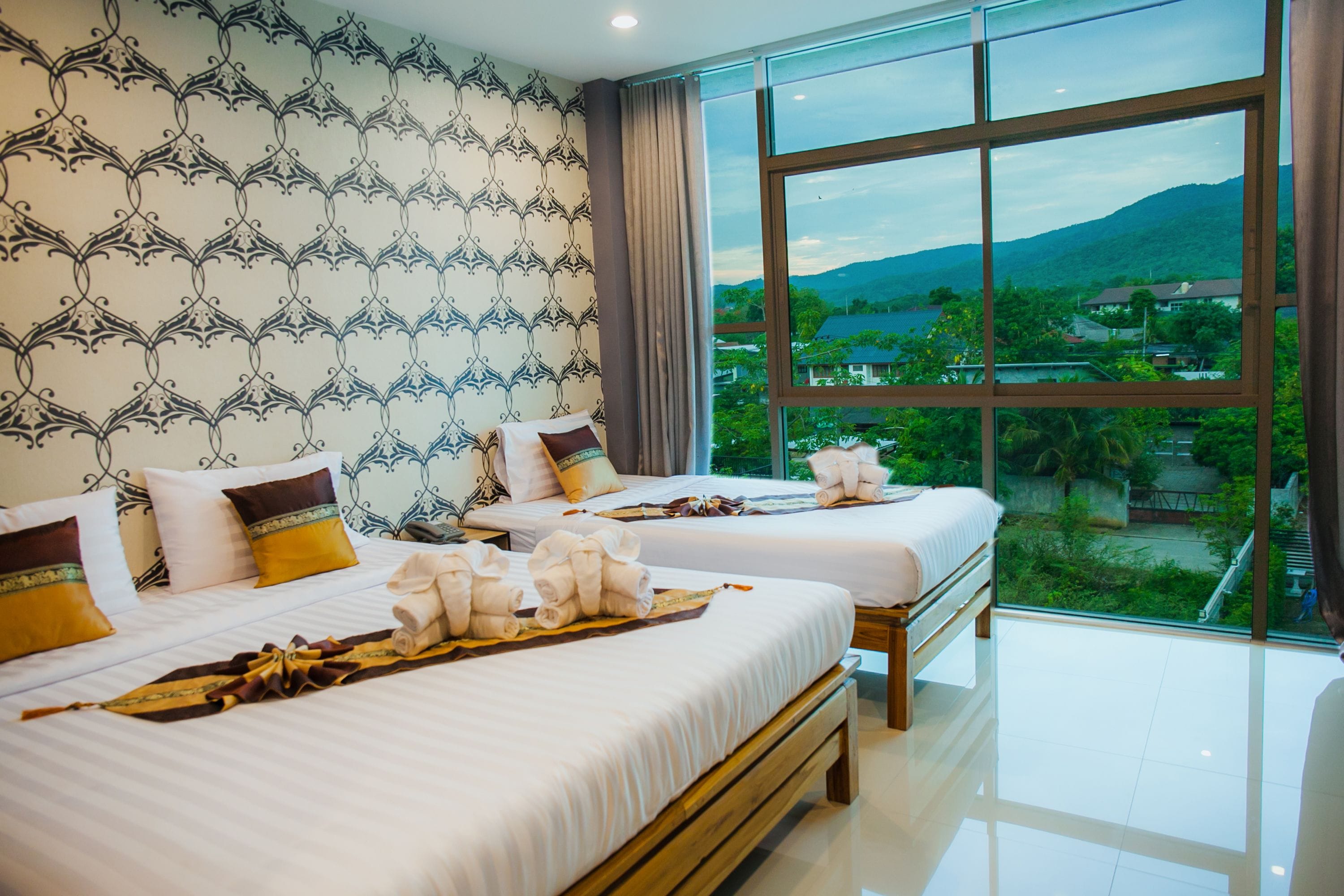 A hotel room with two beds, a large window with a mountain view, and a patterned wallpaper.