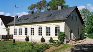 Une maison à deux étages, dotée d’un toit en pente, de nombreuses fenêtres et d’un petit jardin agrémenté de rochers et de plantes.