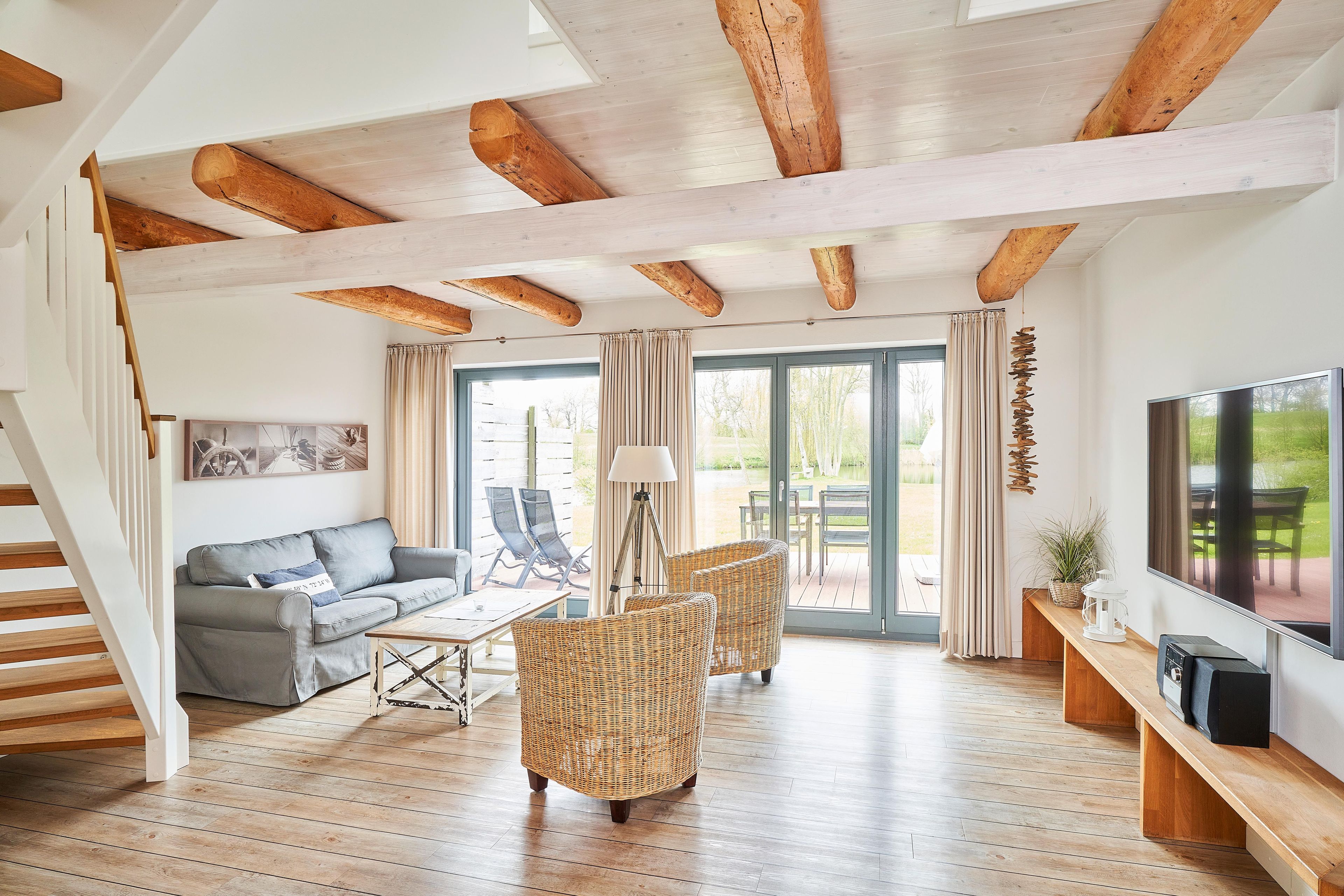 A spacious living area with a wooden ceiling, a grey sofa, wicker chairs, a TV, and a large window with curtains.