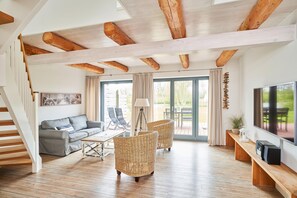 Un vaste espace de vie doté d’un plafond en bois, d’un canapé gris, de fauteuils en osier, d’un téléviseur et d’une grande fenêtre agrémentée de rideaux.