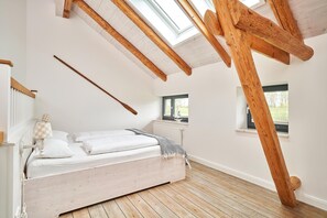 A bedroom with a sloped ceiling, wooden beams, a bed with white linens, a window, and a radiator. - Remise II - Apartments Gut Ostergaard (Steinberg)