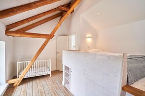 Une pièce aux hauts plafonds avec des poutres apparentes, un berceau blanc, un lit superposé et un miroir au mur.