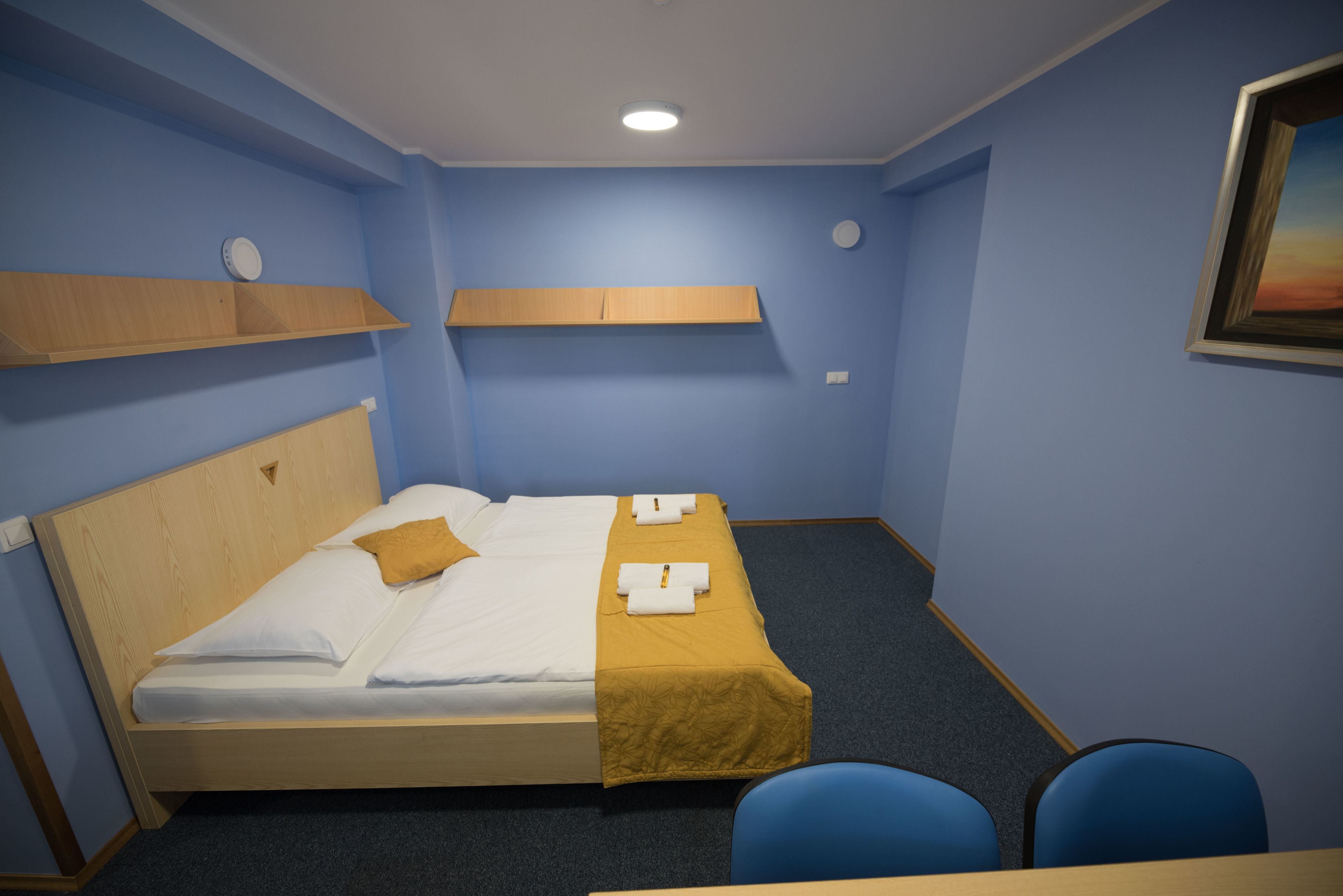 Ein ordentlich bezogenes Bett mit gelber Decke, weißen Kissen, einem hölzernen Kopfteil, einem blauen Teppich und einem kleinen Tisch mit zwei Stühlen.
