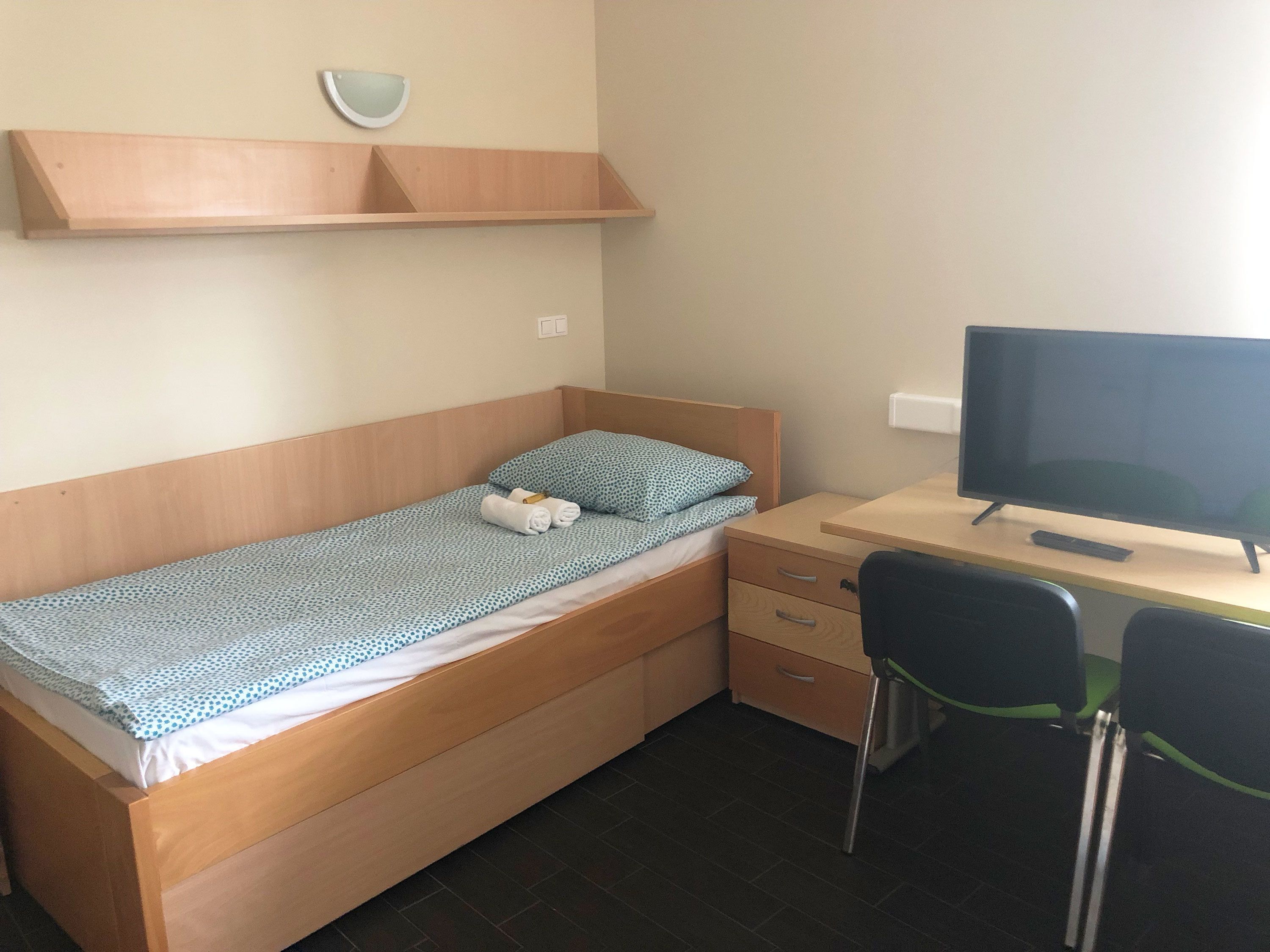 Una camera piccola con un letto singolo, una testiera in legno, una scrivania con televisione e una sedia.