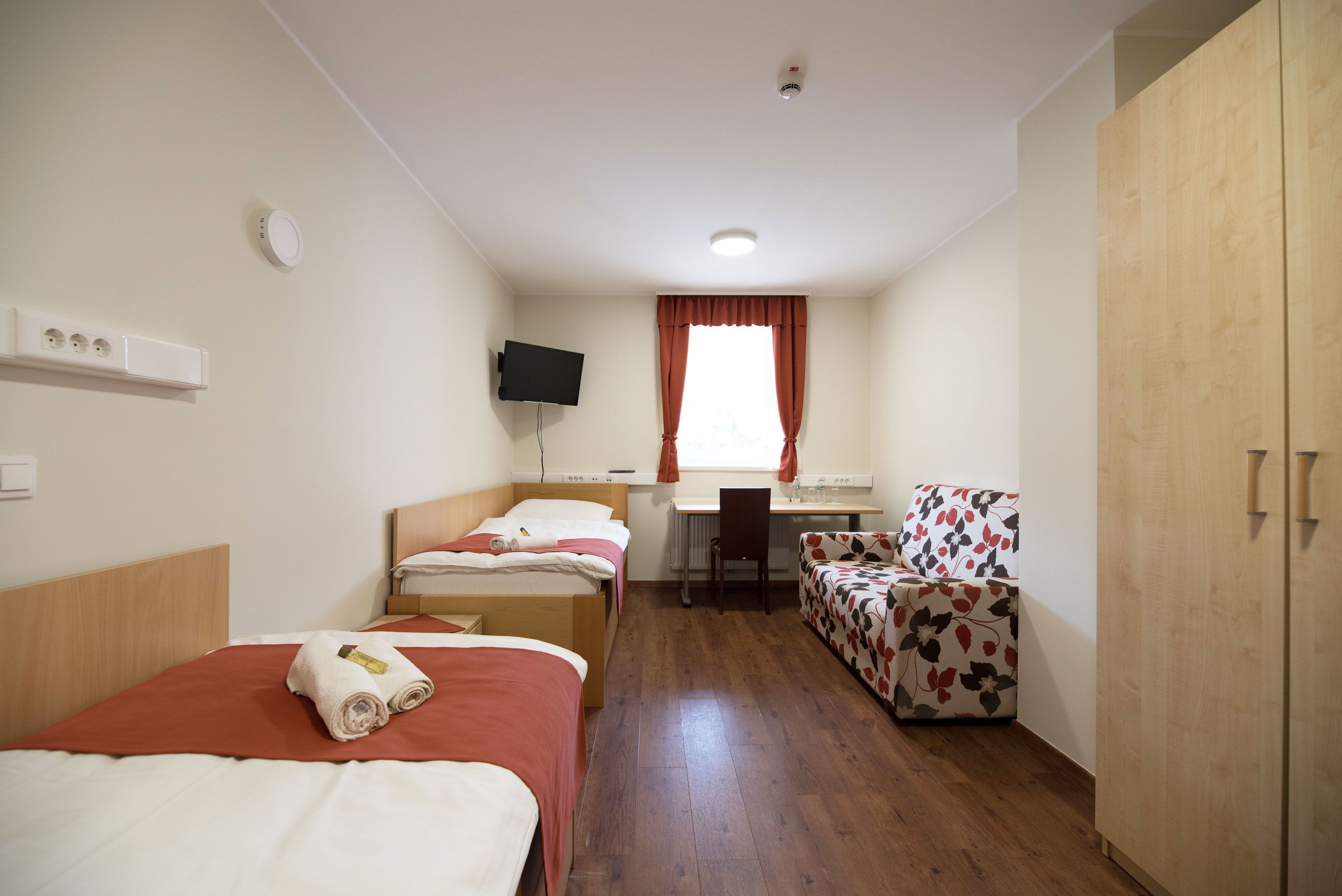 Ein Hotelzimmer mit drei Einzelbetten, einem Schreibtisch mit Stuhl, einem Fernseher und einem Kleiderschrank.