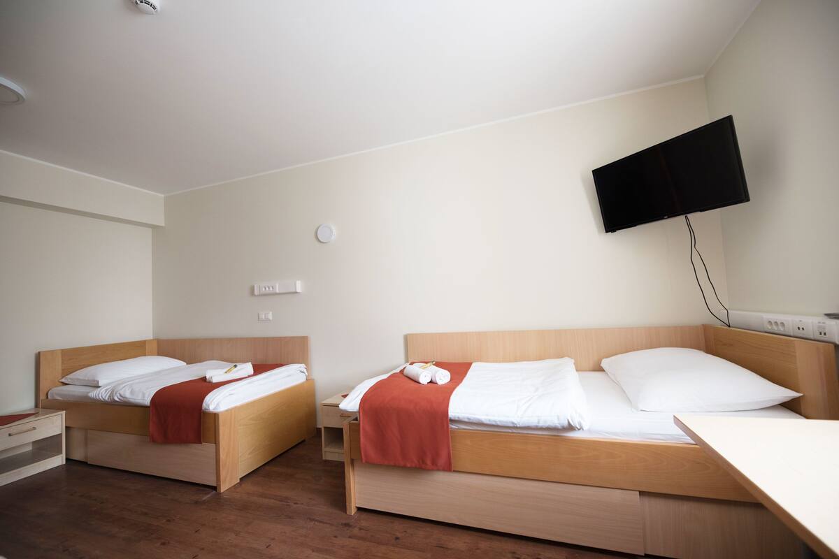 A hotel room with two wooden beds, white bedding, a red blanket, a flat-screen TV mounted on the wall, and a wooden nightstand.