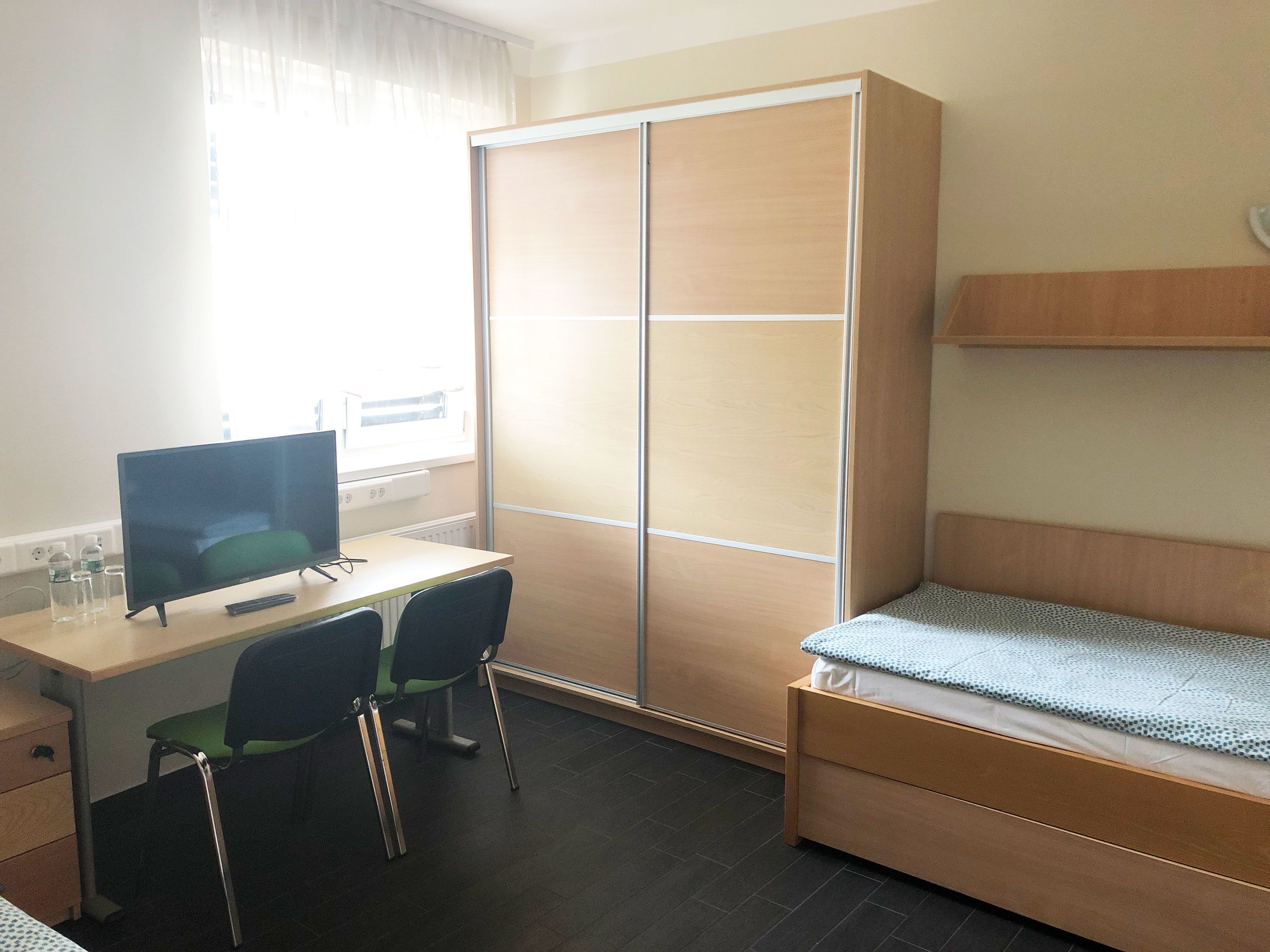 Ein kleiner, gut beleuchteter Raum mit Schreibtisch, zwei Stühlen, einem Bett und einem Kleiderschrank.