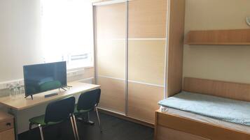 Ein kleiner, gut beleuchteter Raum mit Schreibtisch, zwei Stühlen, einem Bett und einem Kleiderschrank.