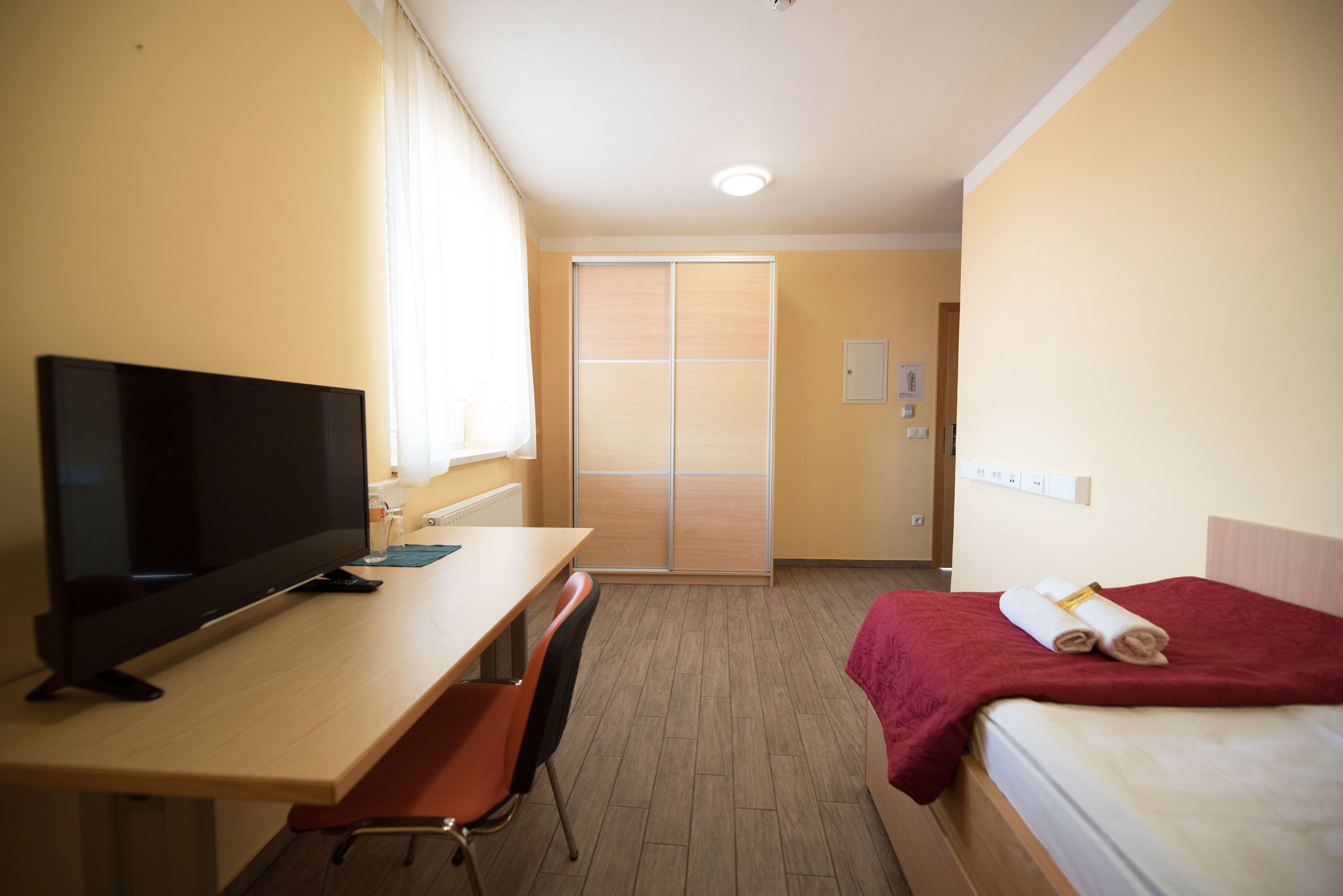 Una habitación de hotel con televisión, escritorio, silla, cama con una manta roja y armario corredero.
