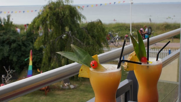 Two cocktails with tropical garnishes on a balcony overlooking a beach with people and a rainbow flag. - Störtebeker Suite, Germany (Glowe)