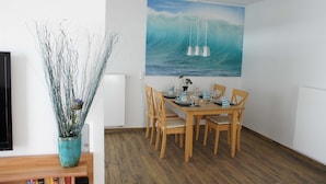 A dining area with a wooden table set for four, a potted plant, and a large wave painting on the wall. - Störtebeker Suite, Germany (Glowe)