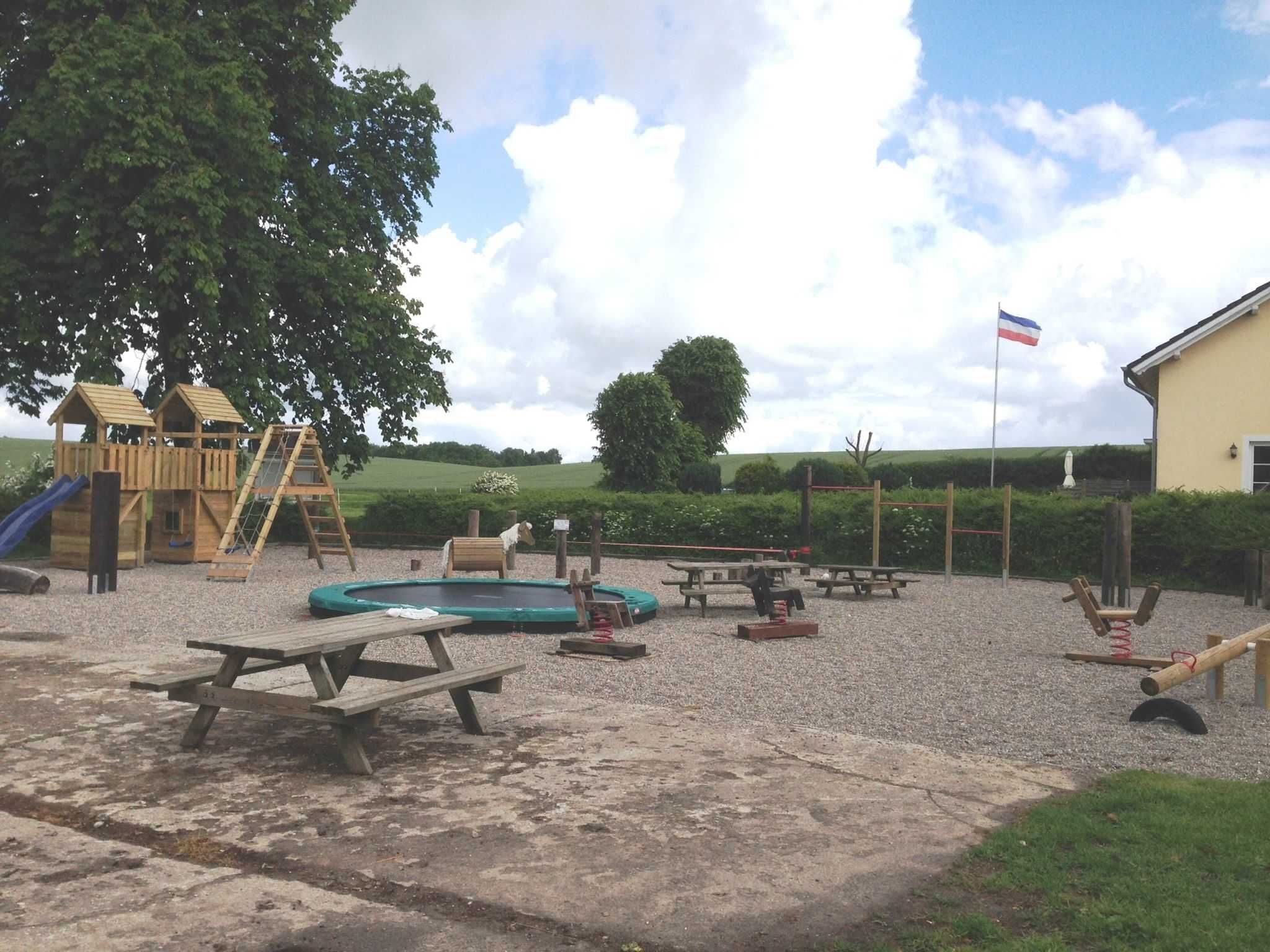 Un parque infantil con estructuras de juego de madera, una piscina pequeña, mesas de picnic y una bandera.