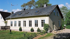 Une maison à deux étages, dotée d’un toit en pente, de nombreuses fenêtres et d’un petit jardin agrémenté de rochers et de plantes.