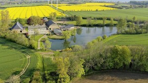Ett lantligt landskap med ett stort byggnadskomplex, en sjö och åkrar med gulgröna grödor.