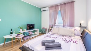 Ein Schlafzimmer mit einem Bett, einem Fernseher, einem kleinen Tisch mit einer Pflanze und einem Fenster mit Vorhängen.