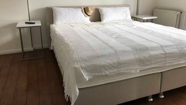 Un lit bien fait, avec une couette blanche et deux oreillers, une table de chevet avec une télécommande, et un radiateur au mur.