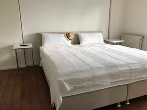 A neatly made bed with a white quilt and two pillows, a bedside table with a remote control, and a radiator on the wall. - Lilly, Glowe on Rügen, Germany (Glowe)