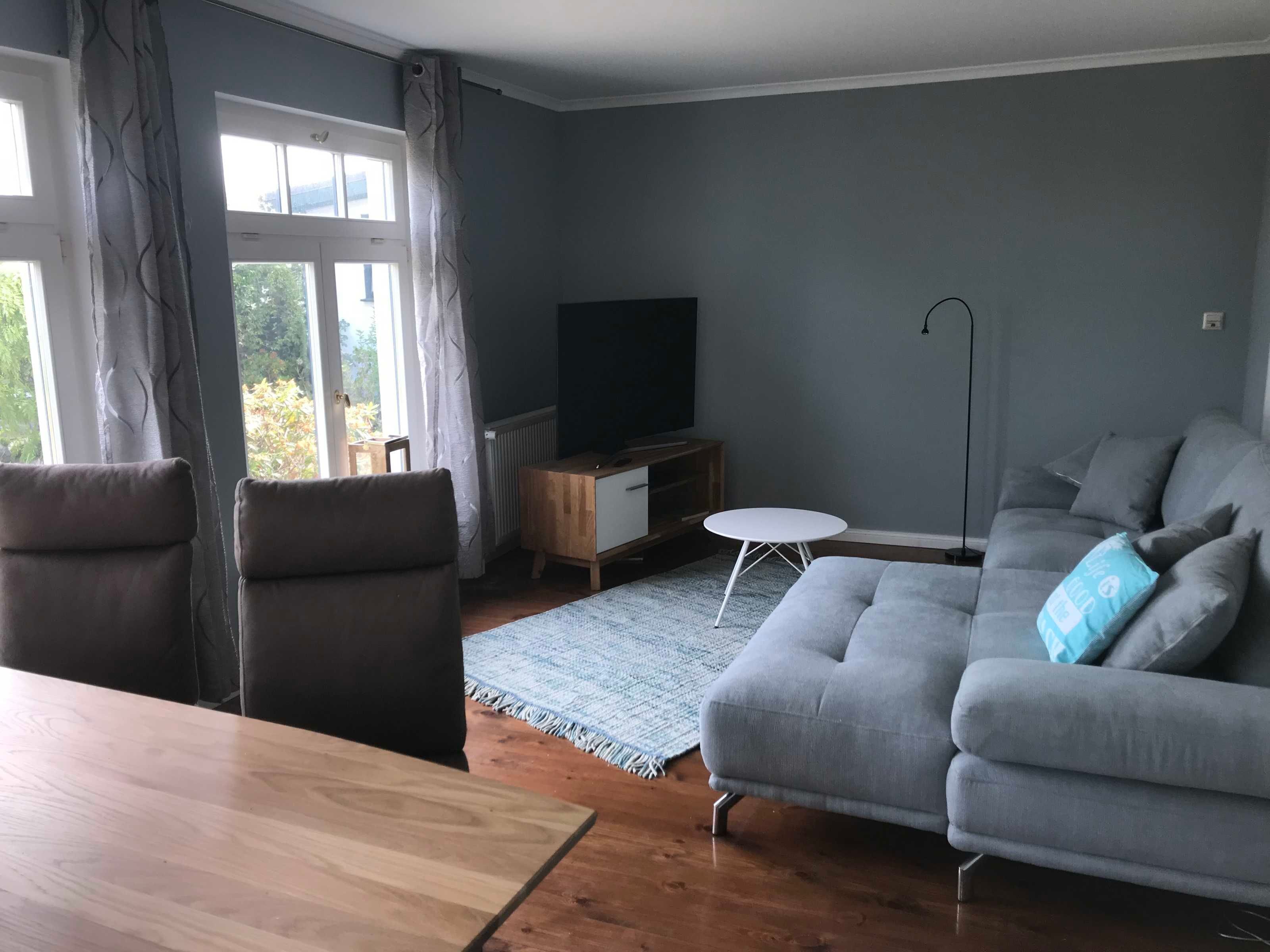 Una sala de estar con un sofá gris, una mesa de comedor de madera y un mueble para televisión.