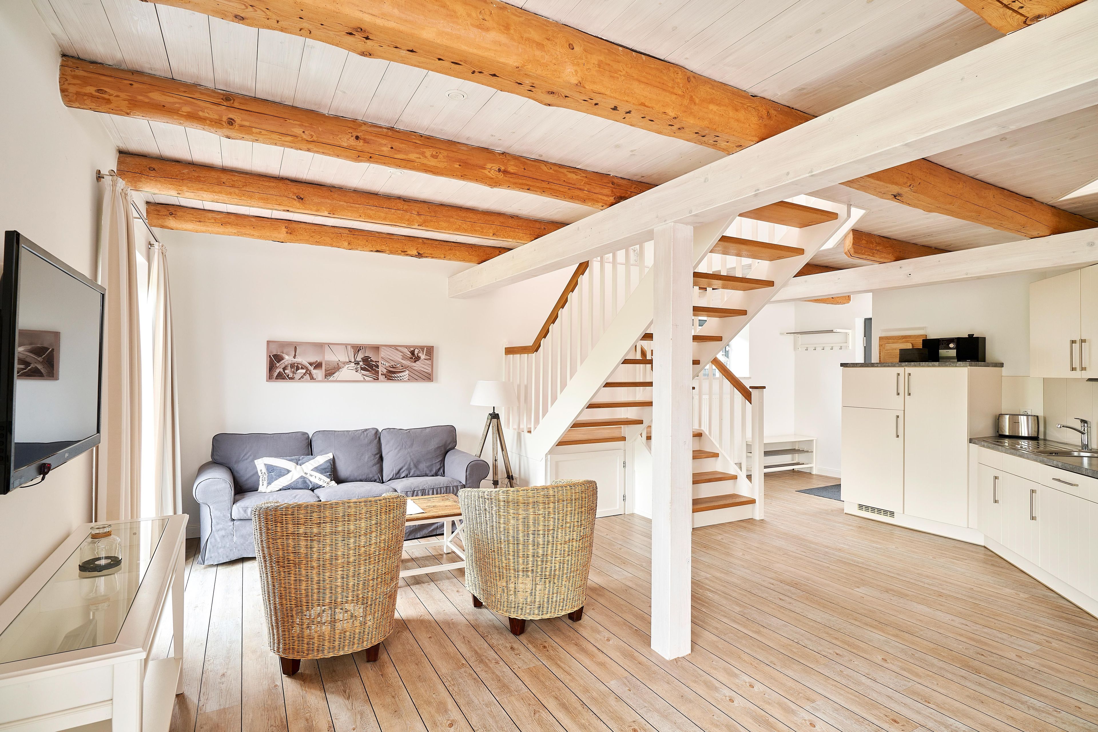A spacious living area with a wooden ceiling, a sofa, wicker chairs, and a staircase leading to a kitchen.