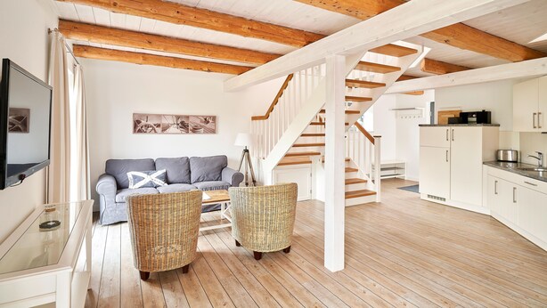 A spacious living area with a wooden ceiling, a sofa, wicker chairs, and a staircase leading to a kitchen. - Remise I - Holiday apartments Gut Oestergaard (Steinberg)