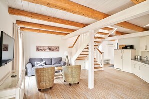 Un vaste espace de vie doté d’un plafond en bois, d’un canapé, de fauteuils en osier et d’un escalier menant à la cuisine.