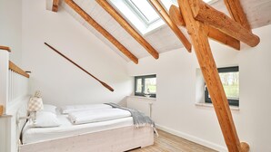 A bedroom with a sloped ceiling, wooden beams, a bed with white linens, a window, and a radiator. - Remise I - Holiday apartments Gut Oestergaard (Steinberg)