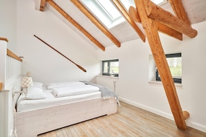 Un dormitorio con techo inclinado, vigas de madera, una cama con ropa de cama blanca, una ventana y un radiador.