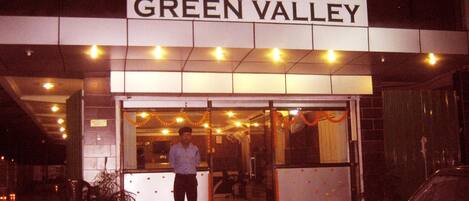 Das Hotel Green Valley mit einem Schild über dem Eingang, einem Mann davor und einem parkenden Auto zur rechten Seite.