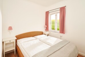 A bedroom with a single bed, a bedside table, a nightstand, a window with curtains, and a lamp on the wall. - Mühlenkate II - Apartments Gut Oestergaard (Steinberg)