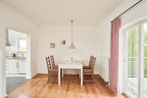 A dining area with a white table and chairs, a hanging pendant light, and a view of the outdoors through a sliding glass door. - Mühlenkate II - Apartments Gut Oestergaard (Steinberg)