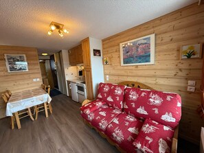 Um quarto acolhedor com piso de madeira, um sofá vermelho com padrão branco, uma mesa de jantar de madeira com cadeiras, um espaço de cozinha compacto com armários de madeira e um lustre.