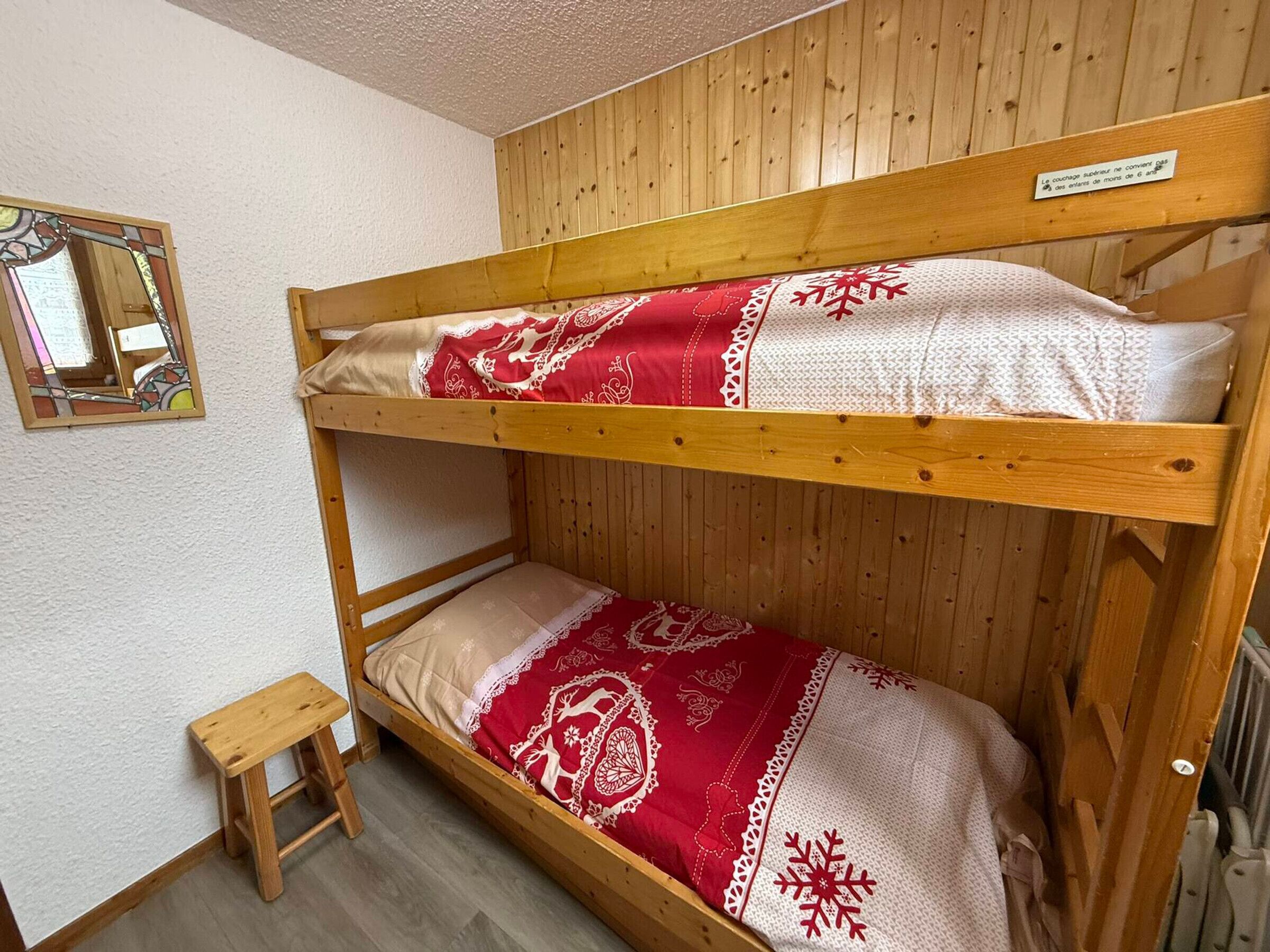 Ein Etagenbett mit rot-weißer Bettwäsche, ein kleiner Holzschemel und ein Spiegel an der Wand.