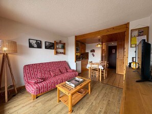 A living room with a red patterned sofa, a wooden coffee table, and a dining area with a wooden table and chairs. - Sunny studio for 4, view on the slopes (Les Saisies)