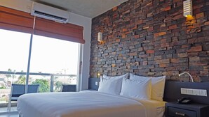 A hotel room with a brick-patterned wall, a large bed with white linens, a bedside table with a phone, and a window with a view of buildings and greenery. - Hotel Sico (Negombo)