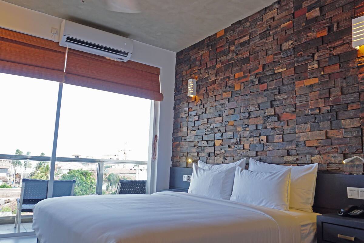 Habitación de hotel con pared de ladrillo, una cama grande con ropa blanca, una mesita de noche con teléfono y ventana con vistas a edificios y follaje.