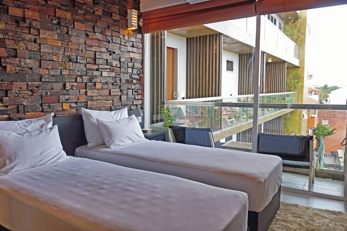 Un dormitorio con dos camas, una pared de piedra y un balcón con vistas a edificios.