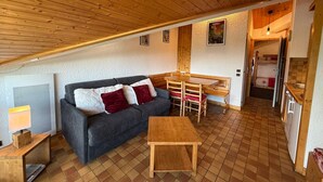 Une pièce chaleureuse avec un plafond en bois, un canapé gris agrémenté de coussins rouges, une table en bois et un petit coin cuisine équipé d’un réfrigérateur et d’un évier.