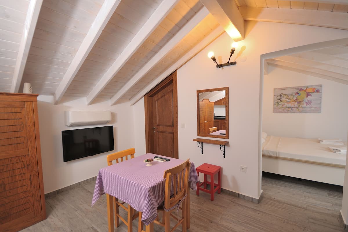 A room with a wooden ceiling, a TV mounted on the wall, a table with a checkered tablecloth, a bed, and a small stool.